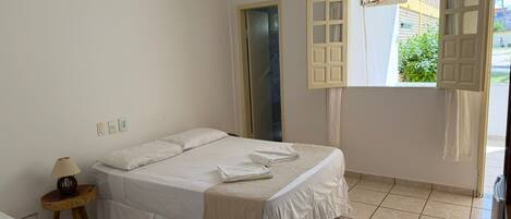 A bedroom with a bed, a bedside table with a lamp, an air conditioning unit, and a window with open shutters.
