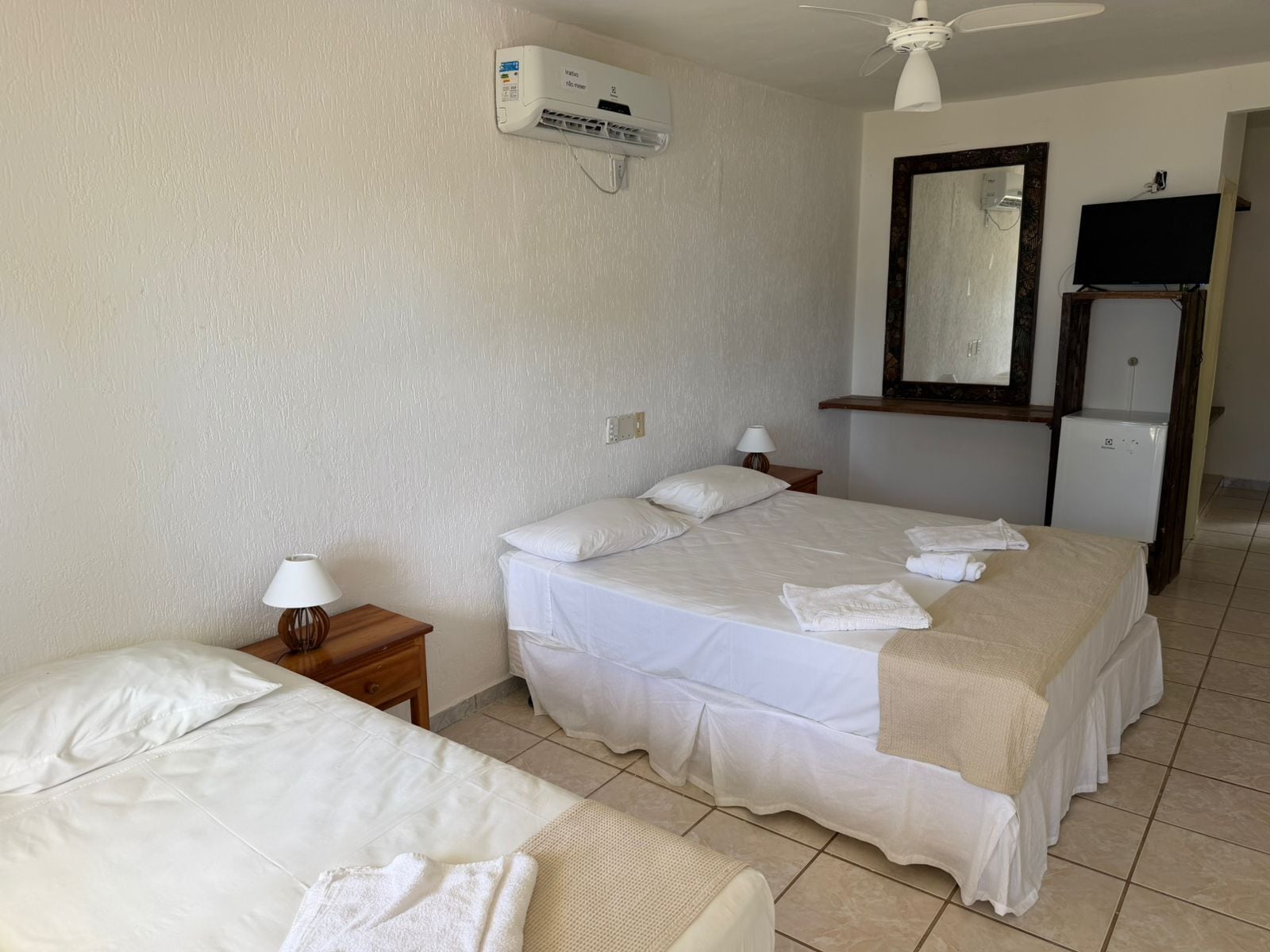 A hotel room with two beds, a ceiling fan, an air conditioning unit, a mirror, a television, and a refrigerator.