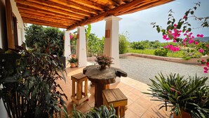 Un espacio al aire libre techado, con techo de madera, plantas en macetas y vistas a un jardín lleno de flores rosadas.