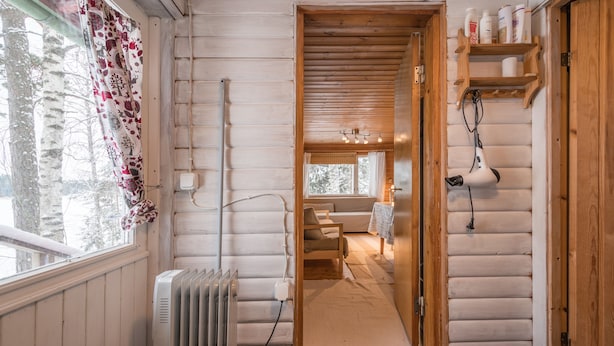 A log cabin interior with a wooden floor, a radiator, a window with curtains, a wooden door, and a shelf with toiletries. - Köningsviken vacations in Sipoo (Sibbo)