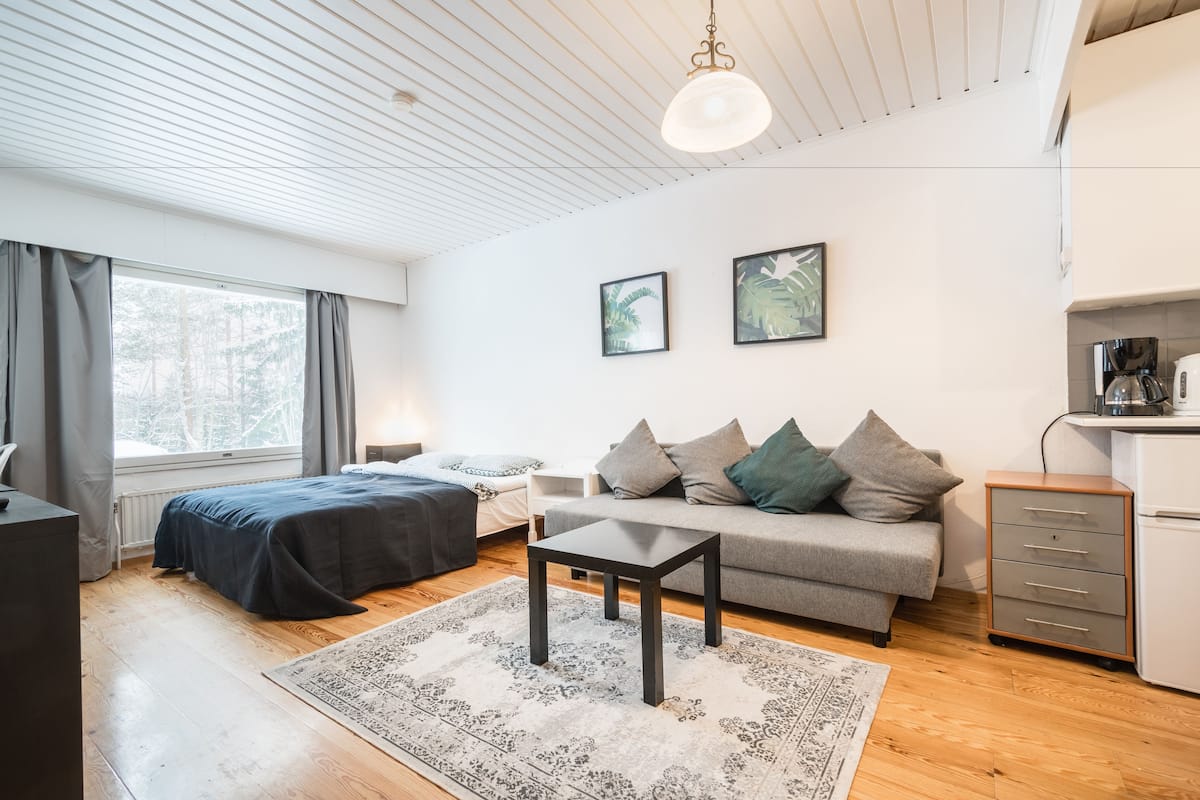 Quarto moderno e bem iluminado com cama, sofá e uma pequena mesa. Há uma janela com cortinas, um teto com ripas de madeira e uma prateleira com chaleira.