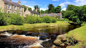 A stone building with multiple chimneys, a small stream flowing through a grassy area, and trees in the background.