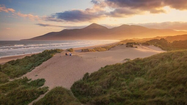 A coastal landscape with a beach, grassy dunes, and a mountain in the background.