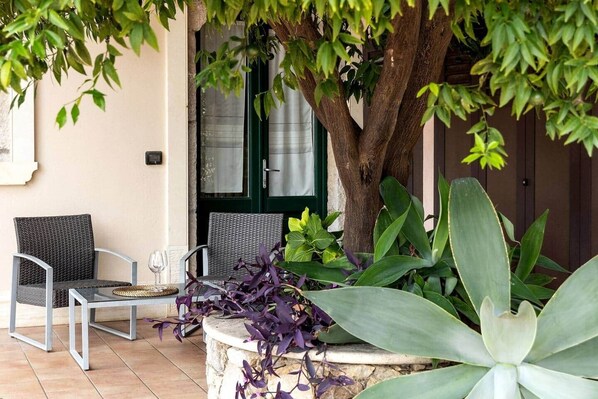 Un espacio al aire libre con dos sillas de mimbre y una mesa, rodeado de plantas verdes grandes y un árbol.
