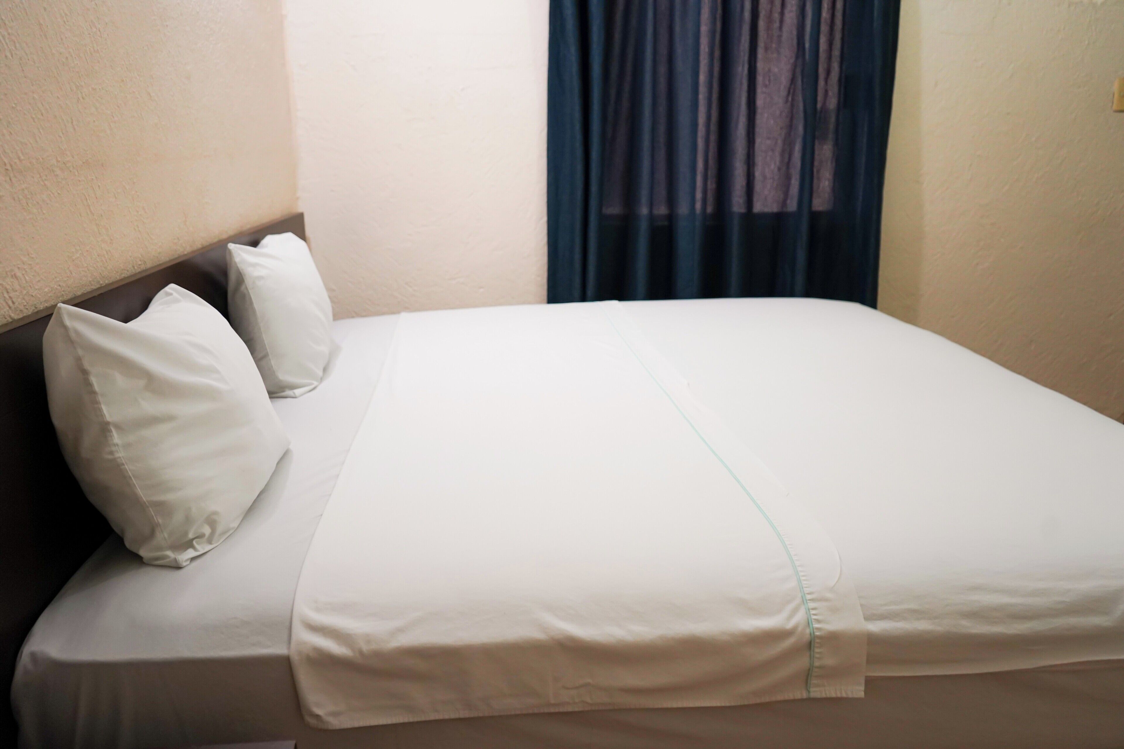 A neatly made bed with white bedding and pillows, a headboard, and a window with dark curtains.