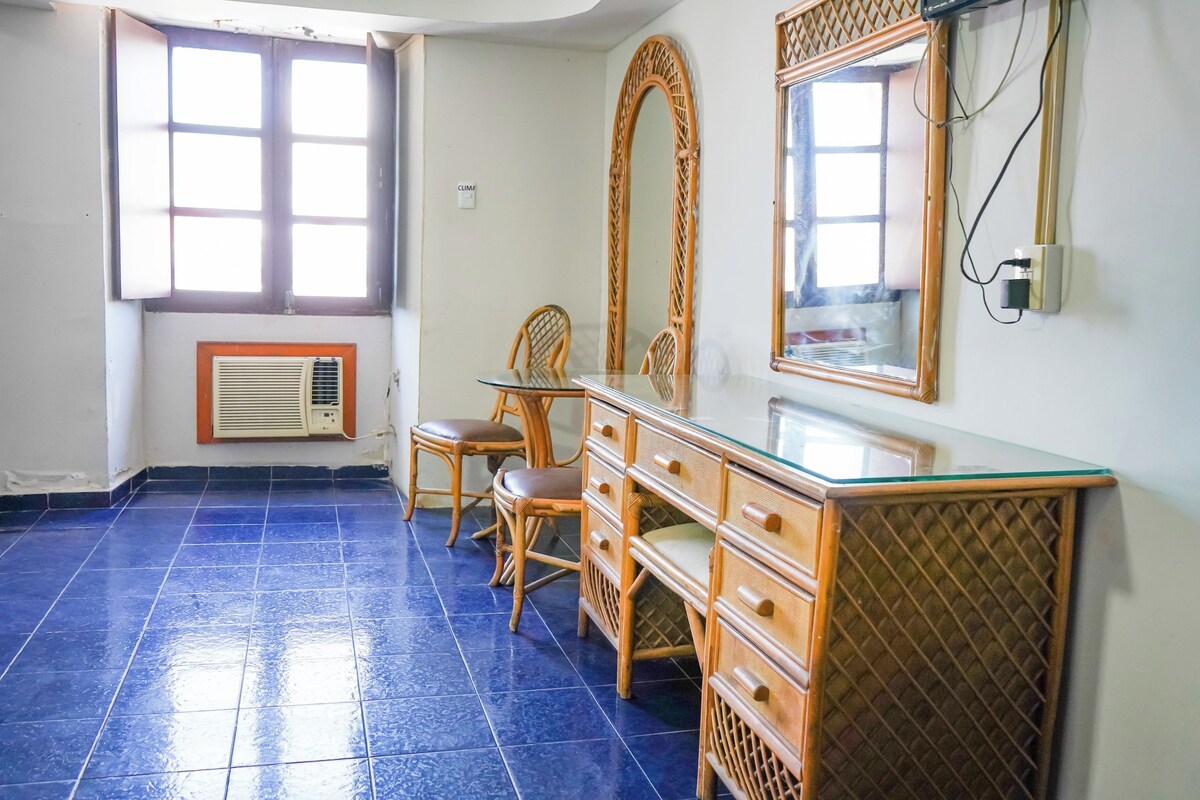 A room with a blue tiled floor, a wooden dresser, two wicker chairs, a large mirror, and a window with a vent.
