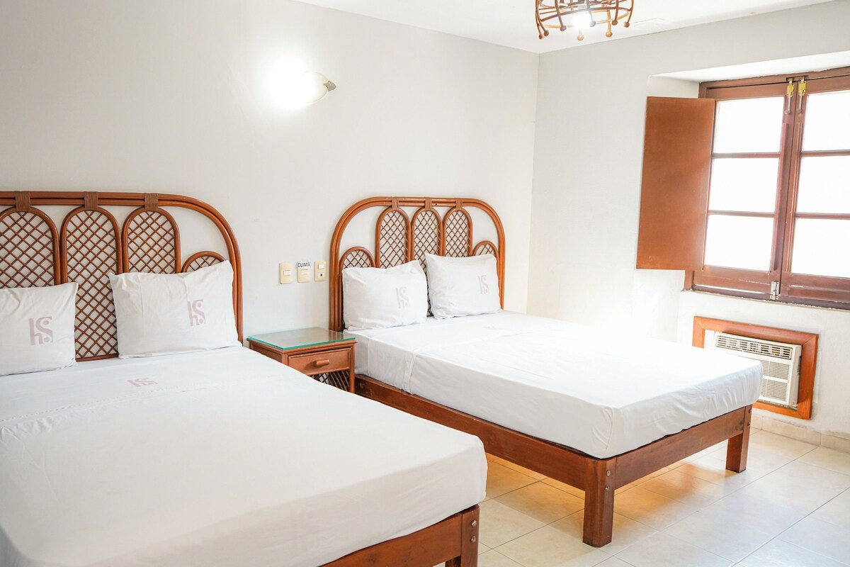 A room with two wooden beds, white bedding, a nightstand, a wall-mounted heater, and a window with a wooden shutter.