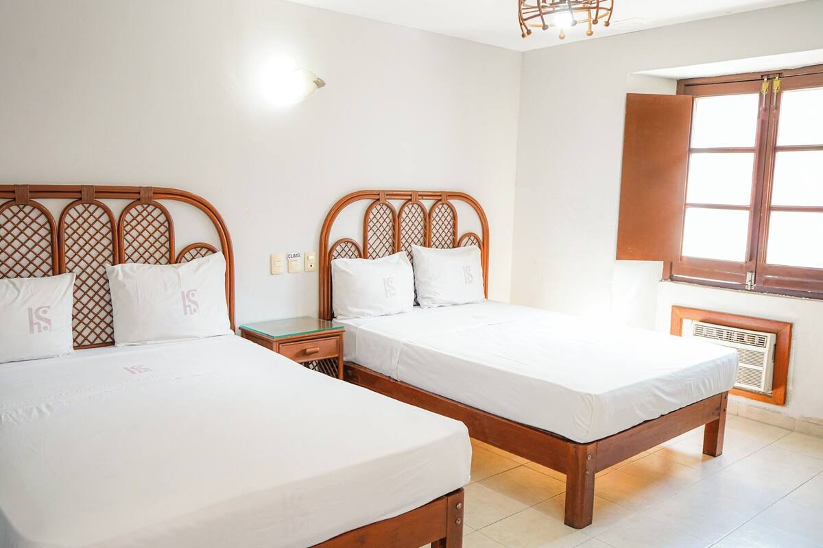 Een kamer met twee houten bedden, witte beddengoed, een nachtkastje, een wandradiator en een raam met houten luik.