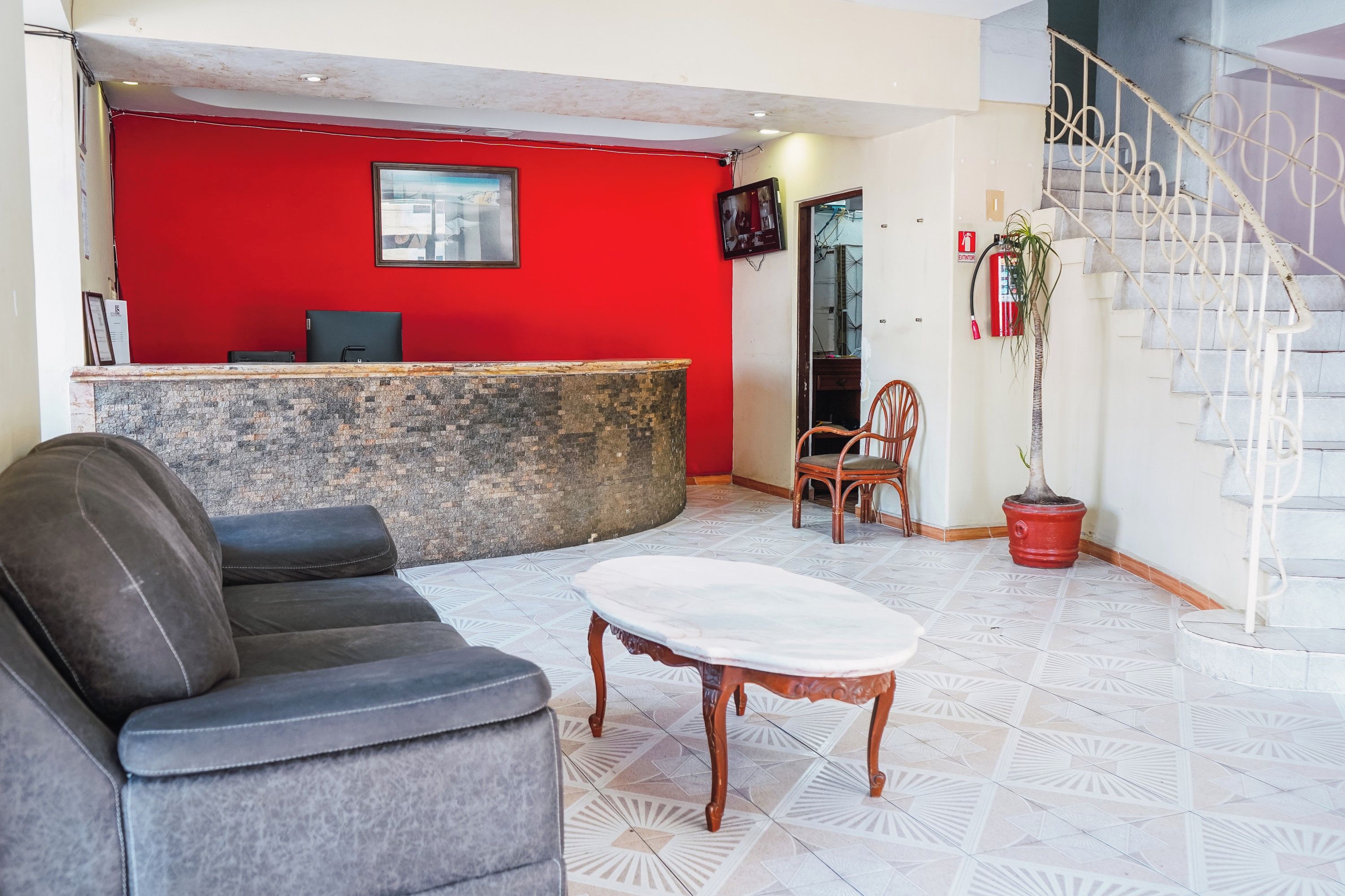 Zona de recepción con pared roja, mostrador, una silla de oficina negra, una silla de madera, una mesa blanca y una escalera con barandilla blanca.
