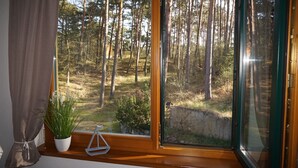 Ein Fenster mit Blick auf einen Wald, eine Topfpflanze und ein kleines, weißes Objekt auf dem Fensterbrett.