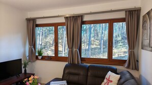 Ein Wohnzimmer mit braunem Ledersofa, einem Flachbildfernseher, einem Couchtisch mit einem Buch und Blumen sowie großen Fenstern mit Vorhängen.