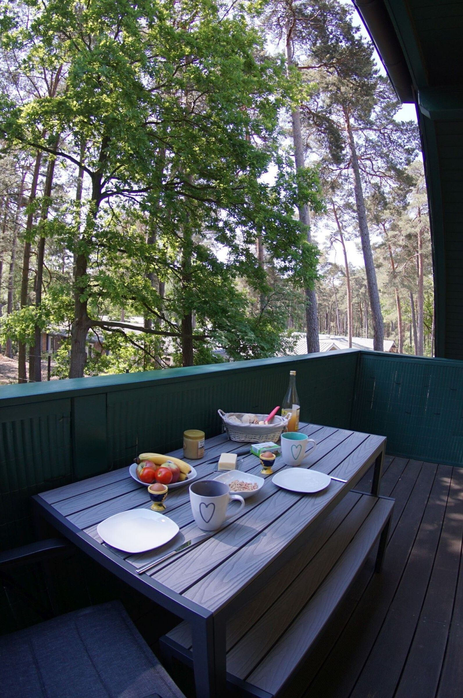 A wooden table with plates, a bowl of fruit, a bottle, and a mug on a balcony with a view of trees.