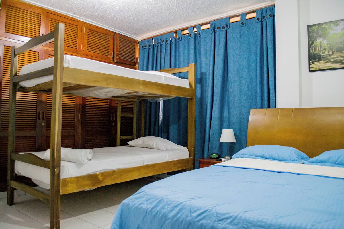 A room with a bunk bed, a single bed, a nightstand with a lamp, and a wall-mounted picture.