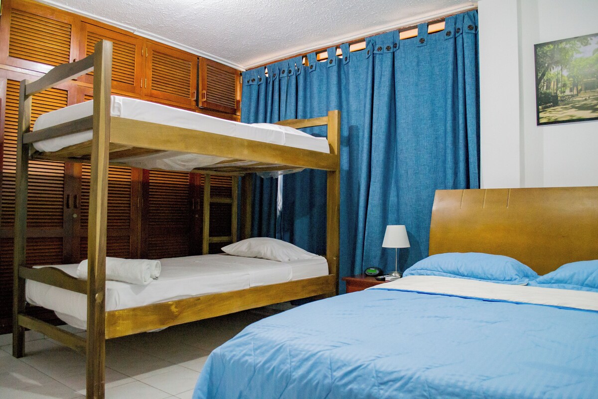 A room with a bunk bed, a single bed, a nightstand with a lamp, and a wall-mounted picture.