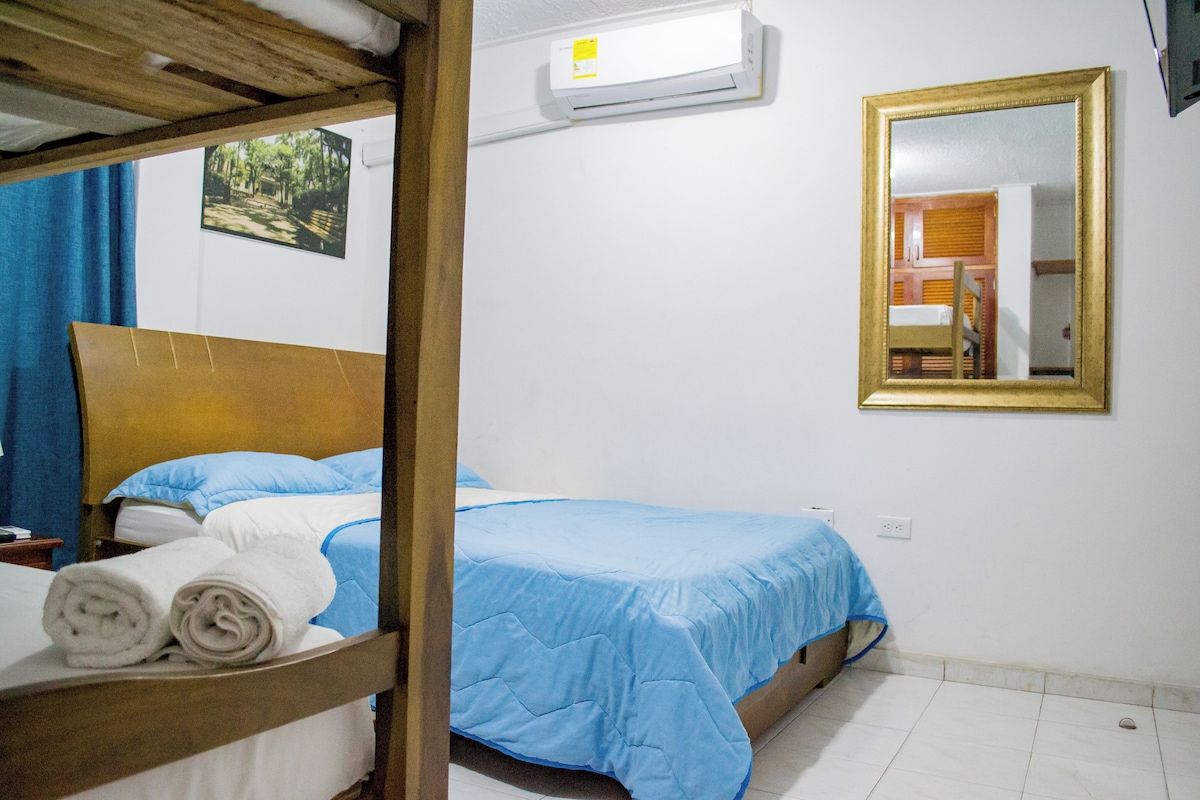 A room with a bunk bed, a single bed with blue bedding, a wall-mounted air conditioner, a mirror, and a picture on the wall.