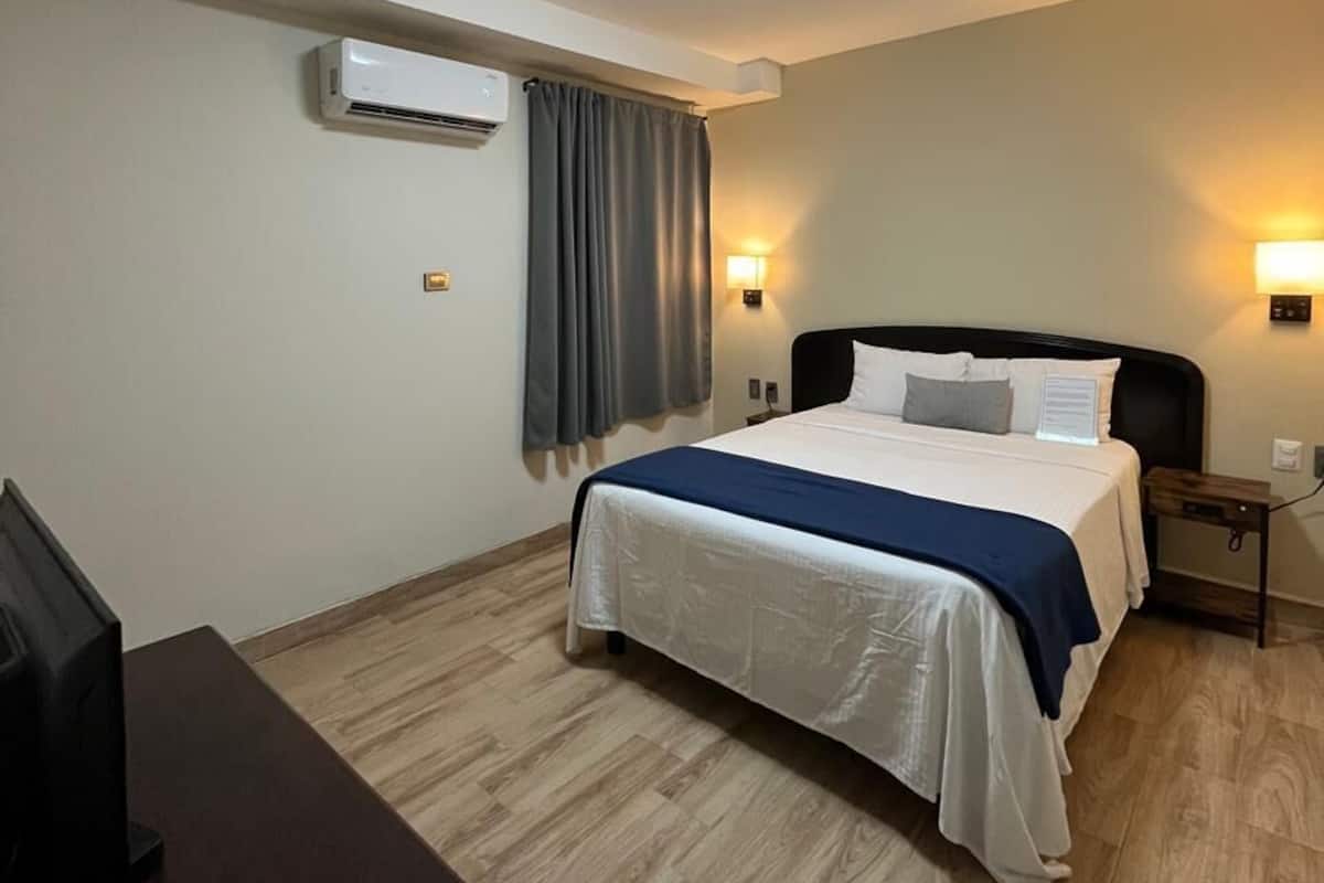 Una habitación de hotel con cama, escritorio, aire acondicionado y una lámpara de pared.