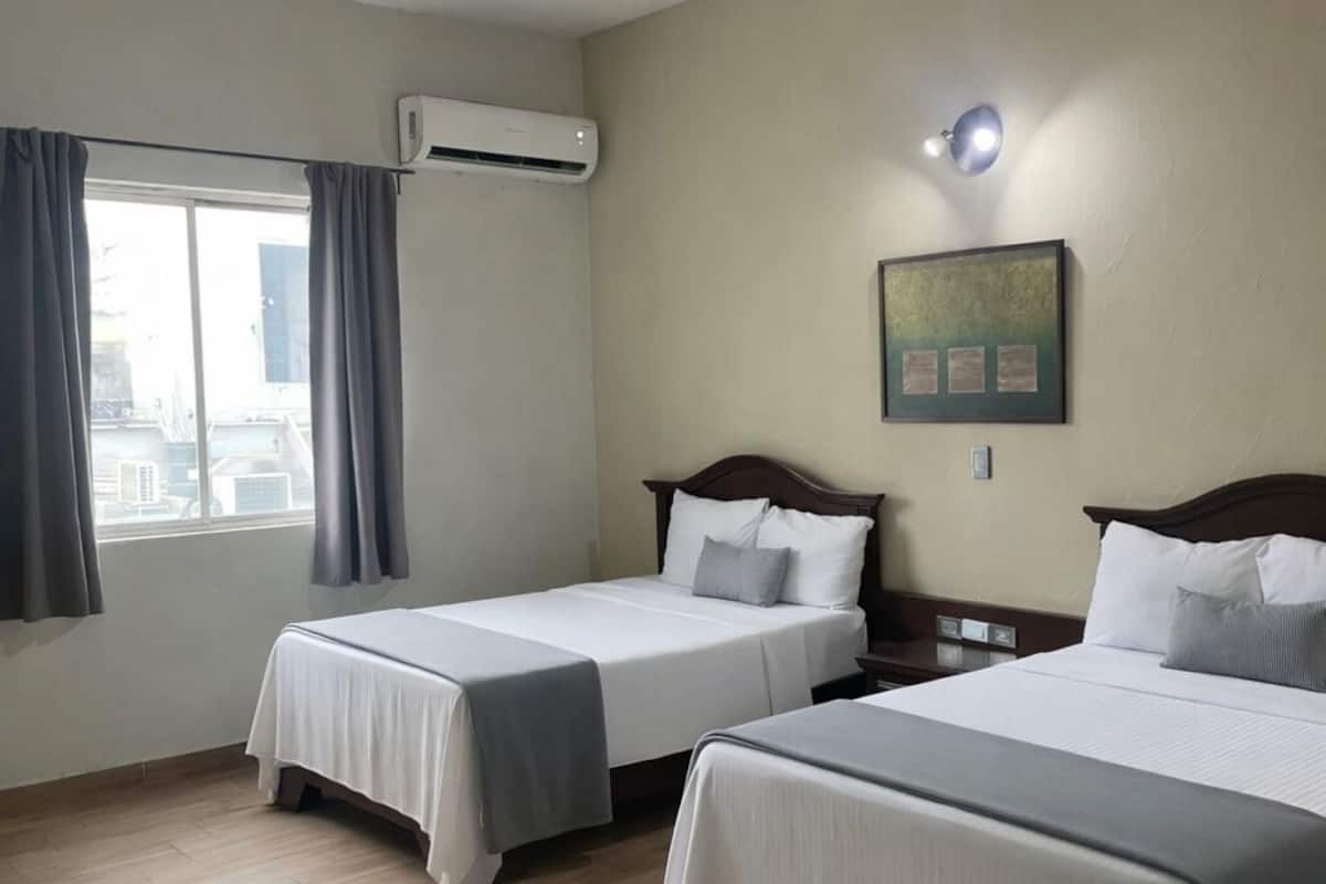 Habitación de hotel con dos camas, una ventana con cortinas, un equipo de aire acondicionado, una lámpara fijada a la pared y un cuadro enmarco.