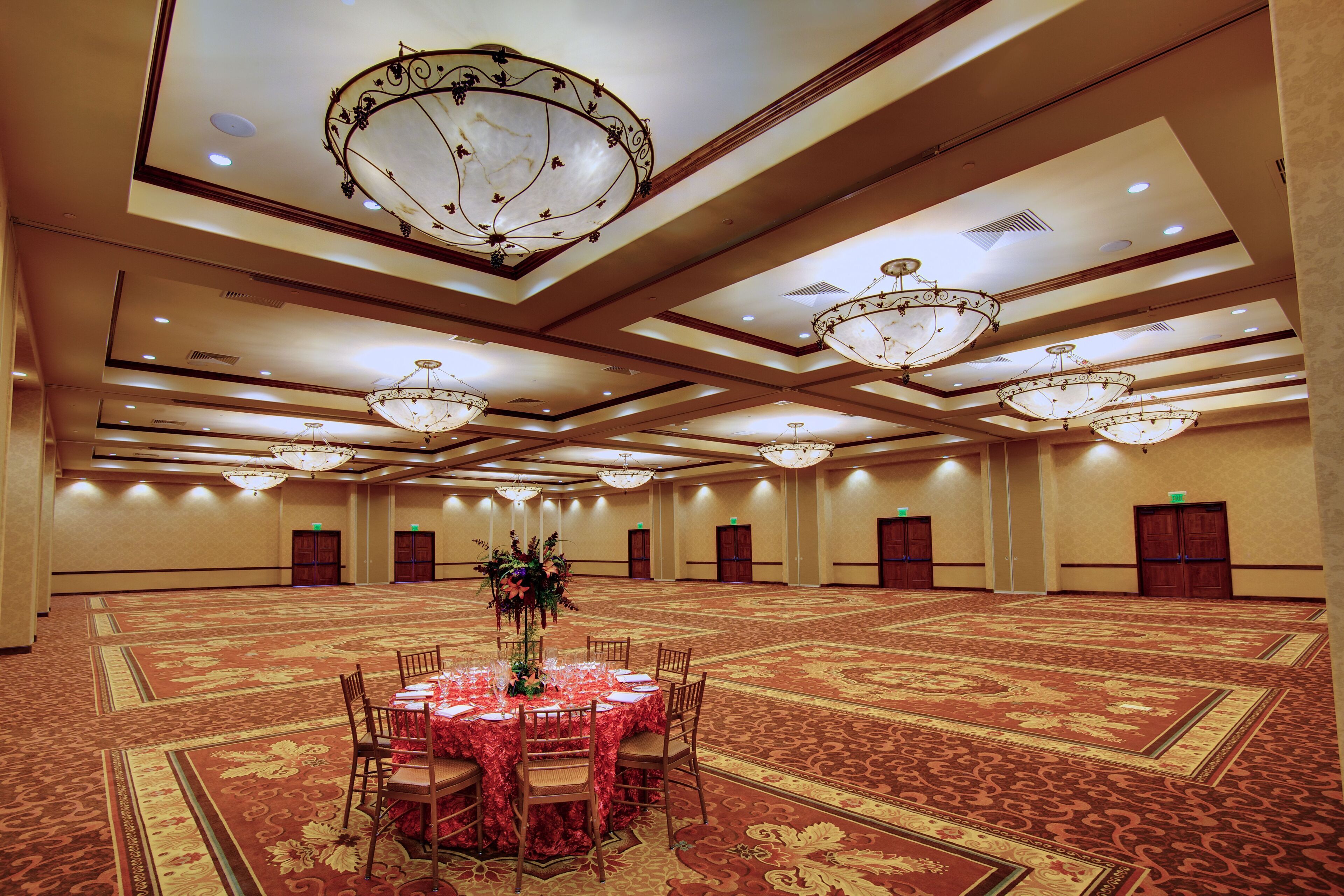 A large, well-lit banquet hall with a central round table set for dining, surrounded by chairs. The room features ornate chandeliers and a patterned carpet.