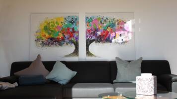 Un salon avec un canapé gris, deux coussins, une table basse en verre et deux tableaux abstraits accrochés au mur.