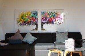 Un salon avec un canapé gris, deux coussins, une table basse en verre et deux tableaux abstraits accrochés au mur.