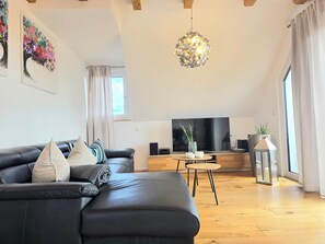 A modern living room with a black leather sofa, a flat-screen TV, a wooden coffee table, and a hanging light fixture.
