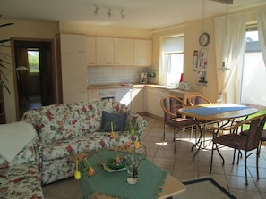 Un salon chaleureux avec un canapé à motifs floraux, une table à manger et des chaises, un coin cuisine doté de placards et d’appareils électroménagers, et une vue sur l’extérieur par une fenêtre.