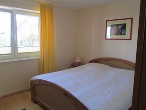 Une chambre avec un lit simple, une fenêtre avec des rideaux jaunes, une table de chevet avec une lampe et un tableau encadré au mur.