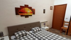 Une chambre à coucher avec une couvre-lit à carreaux, une table de chevet avec une lampe, une œuvre d’art accrochée au mur, une porte et une chaise.