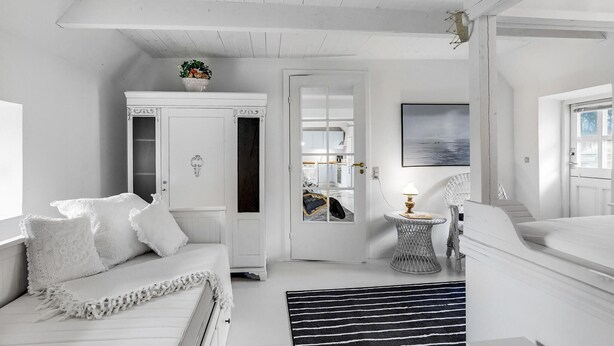 Quarto branco com cama, guarda-roupa, porta que dá para uma cozinha e uma pequena mesa com abajur.