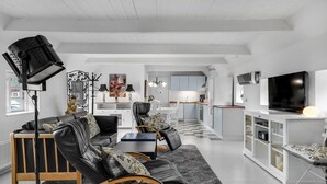 Uma sala de estar moderna com um sofá, poltronas e uma mesa de centro. Uma cozinha visível ao fundo, com armários brancos e um espaço de jantar.