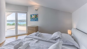 Un dormitorio con cama, mesita de noche, un cuadro y vista al mar a través de un ventanal de vidrio.