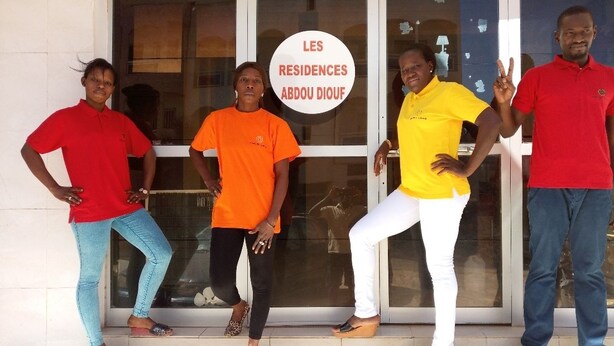 Quatro pessoas em frente a um edifício com a placa "LES RESIDENCES ABDOU DIOUF".