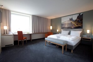A hotel room with a bed, desk, two chairs, a window with curtains, a painting on the wall, and a nightstand with a lamp.