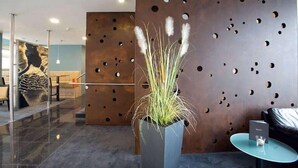 Un área de recepción moderna con una pared de metal que presenta círculos recortados, una planta en maceta con flores blancas y una pequeña mesa con un vaso.