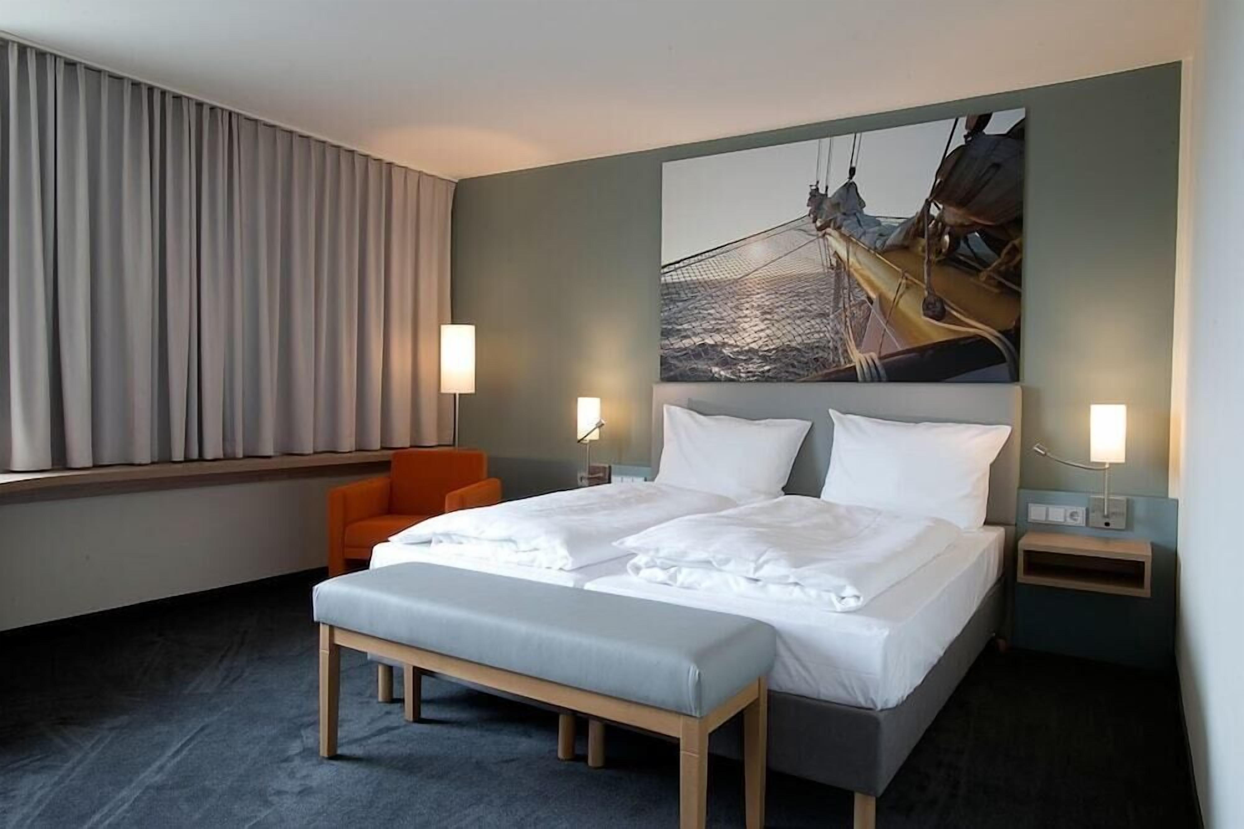 Ein Hotelzimmer mit einem großen Bett, zwei Nachttischen, einer Bank und einem orangefarbenen Sessel, sowie einem großen Gemälde an der Wand.