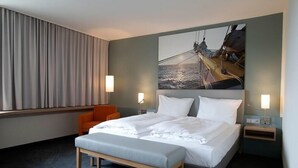 Habitación de hotel con una cama grande, dos mesitas de noche, un banco, una silla naranja y un cuadro grande en la pared.