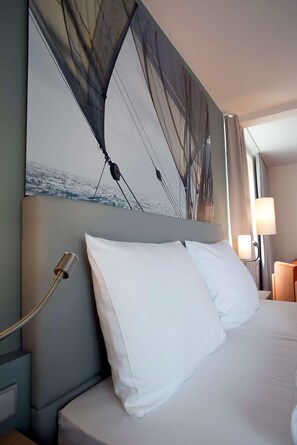Une chambre d’hôtel avec un grand lit, des oreillers blancs, une tête de lit ornée d’un tableau, une lampe de chevet et une table de chevet.