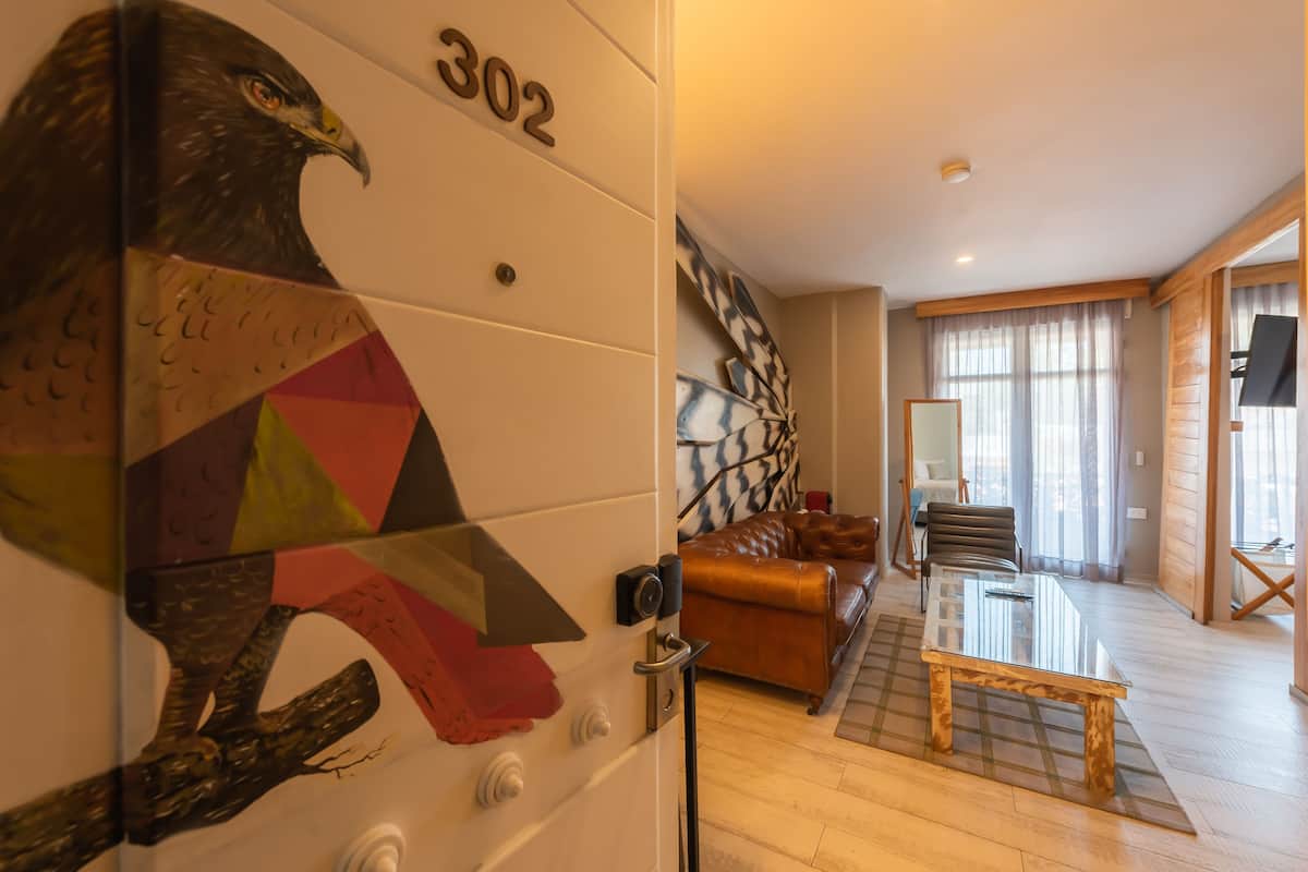 Une chambre d’hôtel avec une grande fresque murale représentant un aigle, un plancher en bois, un canapé en cuir, une table en verre et une grande fenêtre avec des rideaux.