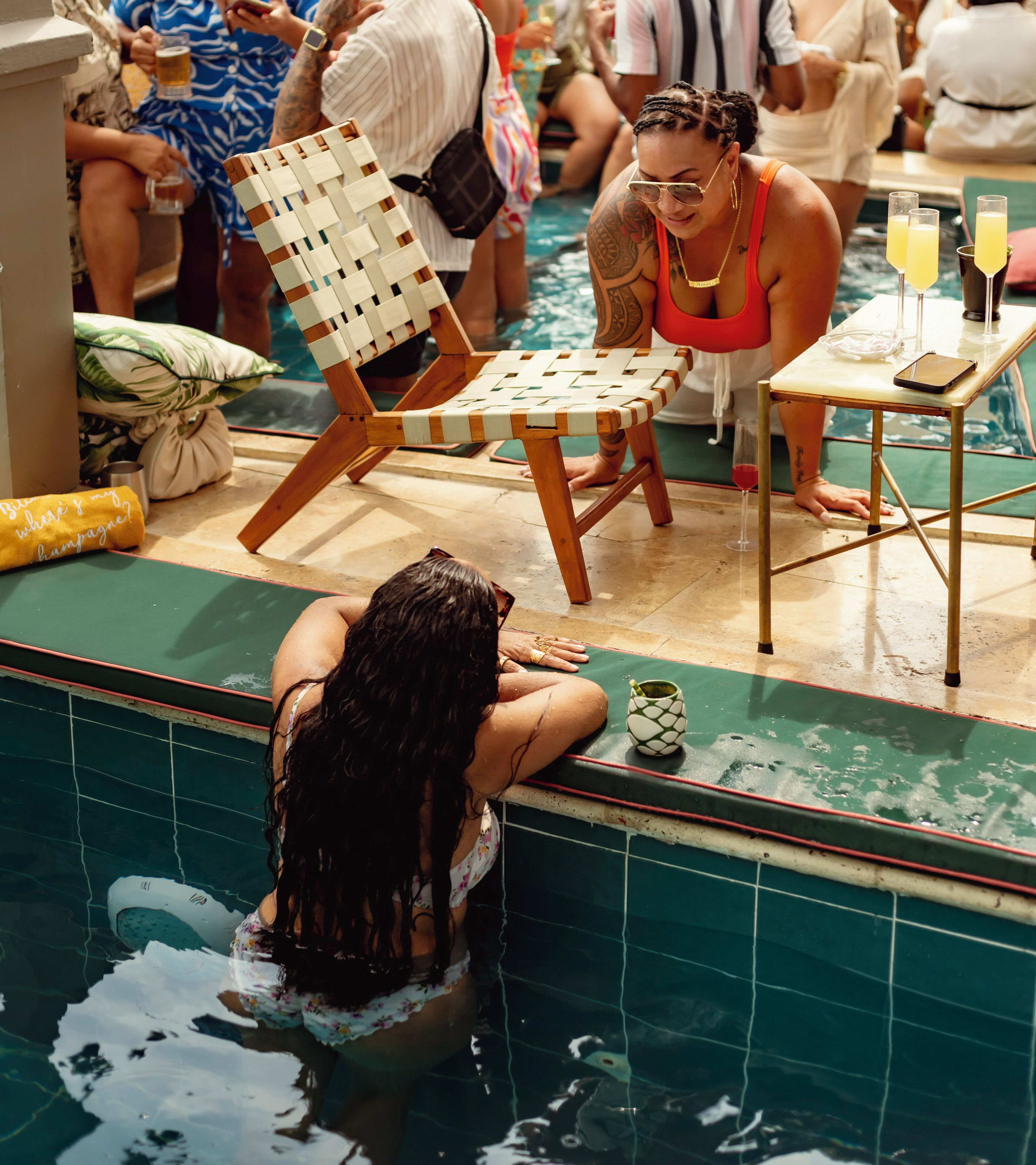 A poolside area with a lounge chair, a table with drinks, and a person leaning on the pool edge.