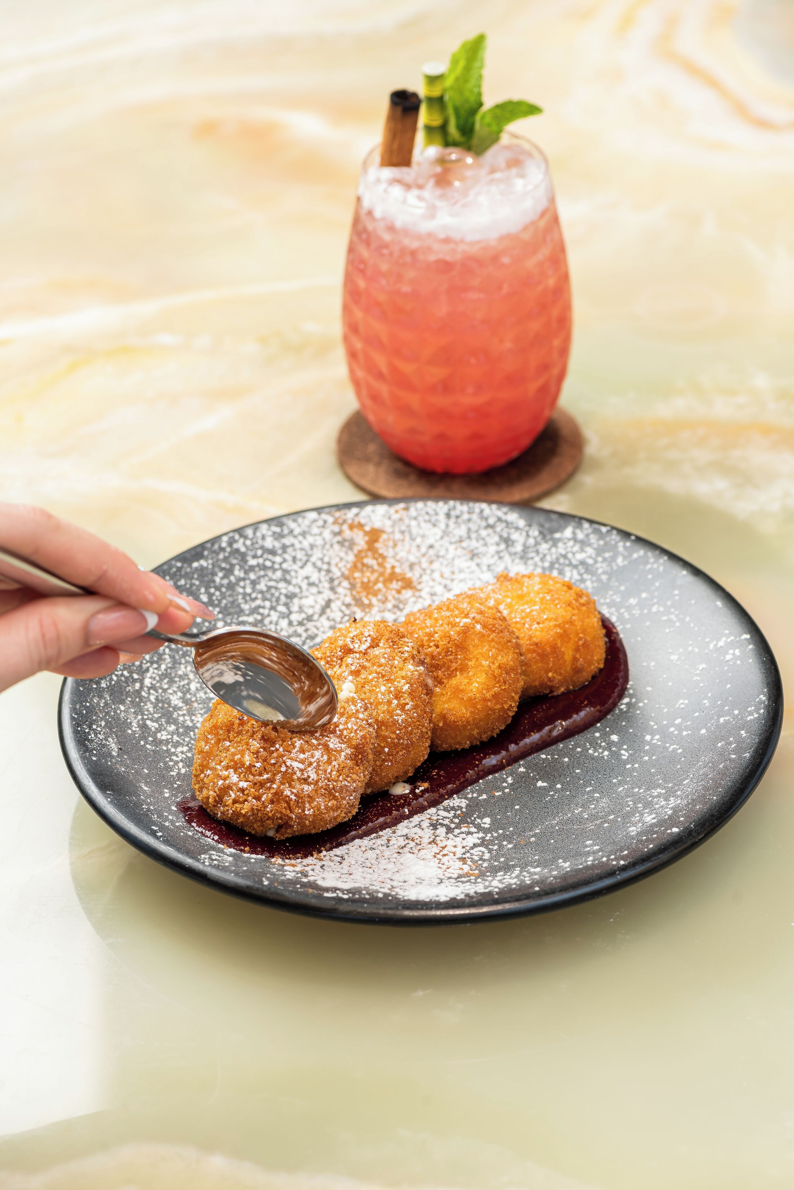 Un plato de comida empanizada y frita, espolvoreada con azúcar glas, una cuchara y un vaso de bebida rosa con adorno de menta sobre una superficie de mármol.