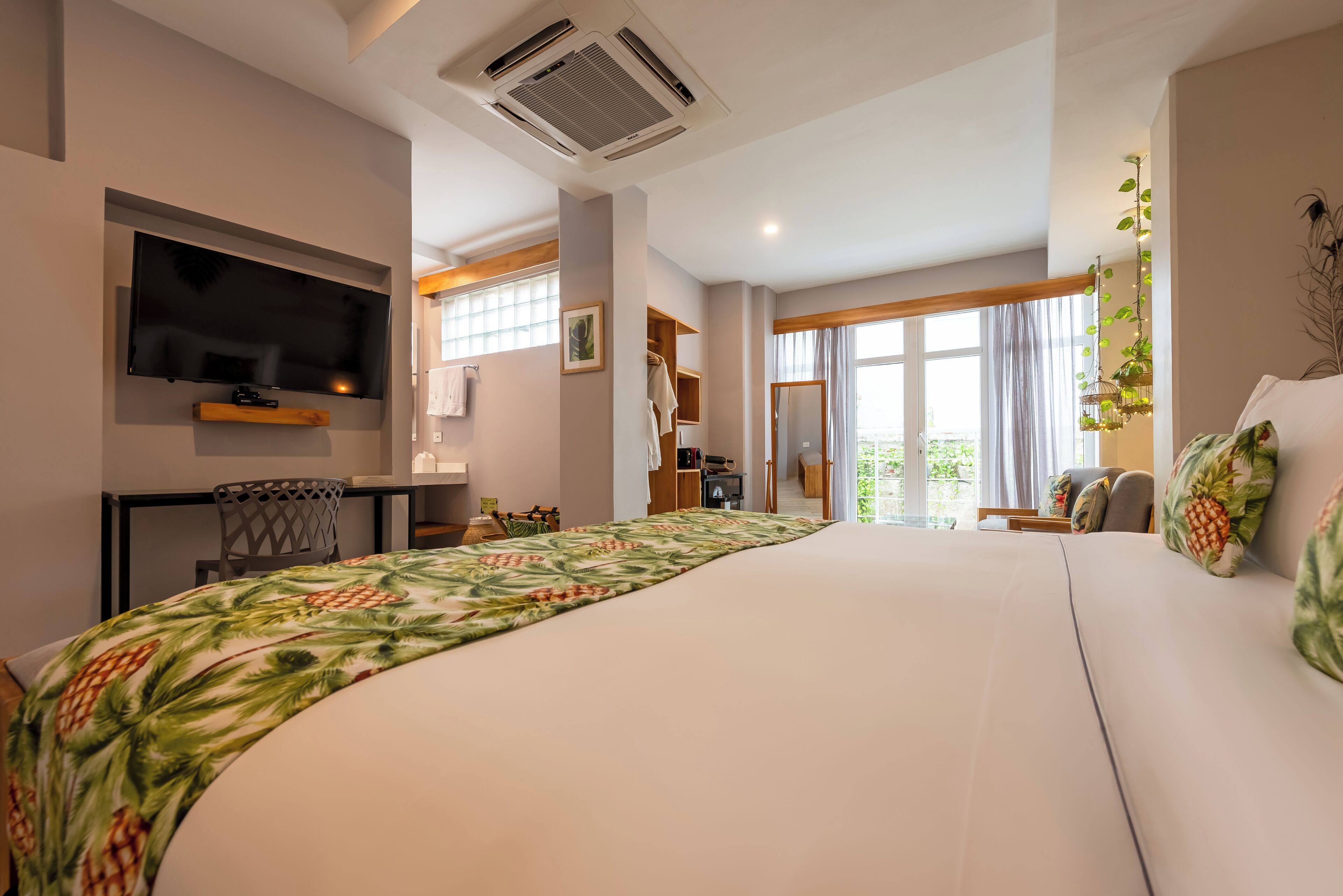 A spacious hotel room with a large bed, a flat-screen TV mounted on the wall, a wooden desk, and a view of the outdoors through a large window.