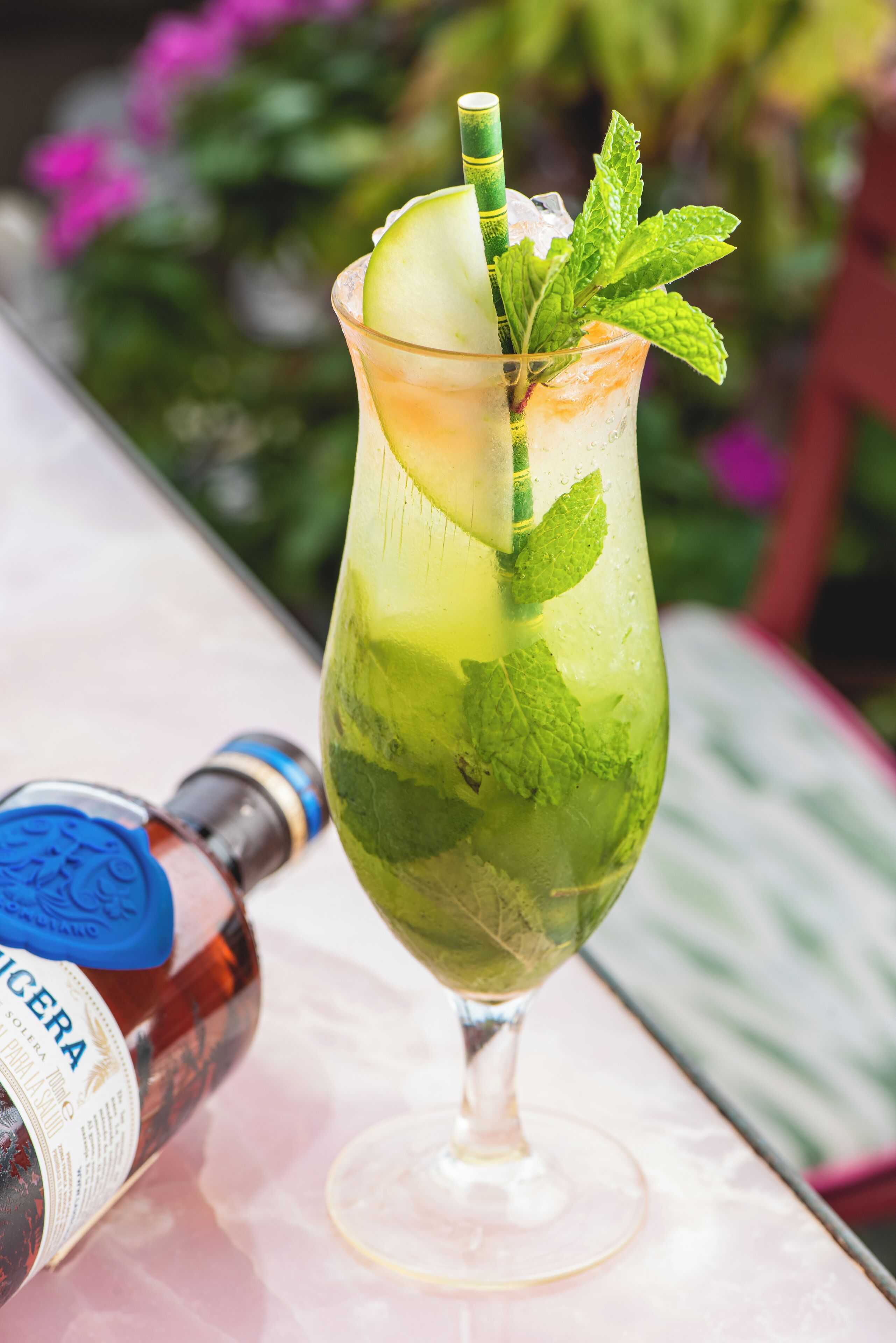 Un cóctel en un vaso con una cuña de lima y hojas de menta, una botella de licor y una mesa con superficie rosa.