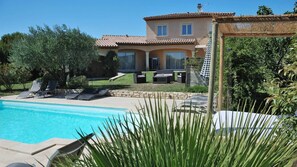 Una piscina con sillones de descanso, una casa con techo de tejas y un jardín con diversas plantas.