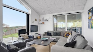 A modern living room with a grey sofa, a black leather chair, and a coffee table. There is a flat-screen TV mounted on the wall, and a large window providing a view of the outdoors. - Nice home in Glesborg with sauna (Glesborg)