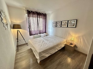 A bedroom with a bed, a desk, a lamp, a window with curtains, and framed pictures on the wall. - Charming Apartment in Cap d'Agde, 2 Bedrooms, Pool, Garage, Close to Beach (Cap d'Agde)