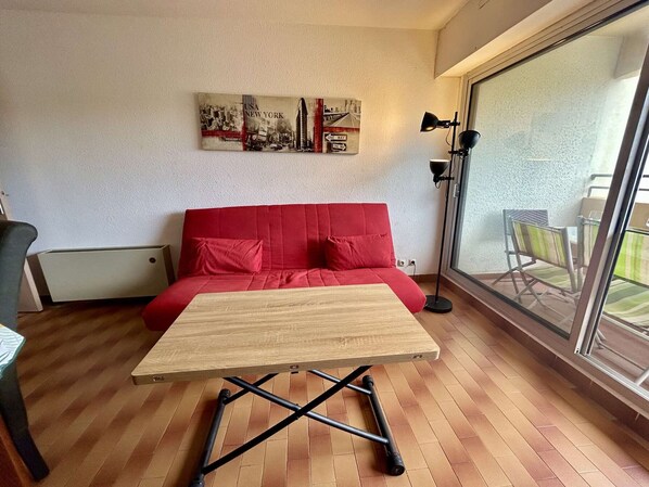 A room with a red sofa, a wooden table, a floor lamp, and a cityscape artwork on the wall. - Charming Apartment in Cap d'Agde, 2 Bedrooms, Pool, Garage, Close to Beach (Cap d'Agde)