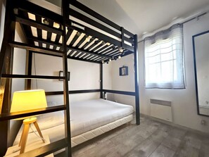 A bunk bed room with a wooden frame, a bedside lamp, a small stool, a window with curtains, and a radiator.