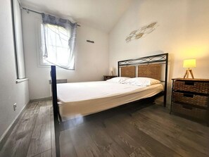 A single bed with a metal frame and a headboard with woven panels, a bedside table with a lamp, and a window with a curtain.