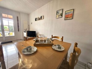 A dining area with a wooden table set for two, a television in the background, and a window with a view of the outside.