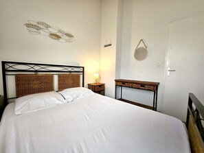 A bed with a woven headboard, a bedside table with a lamp, a small mirror, and a wicker storage unit.